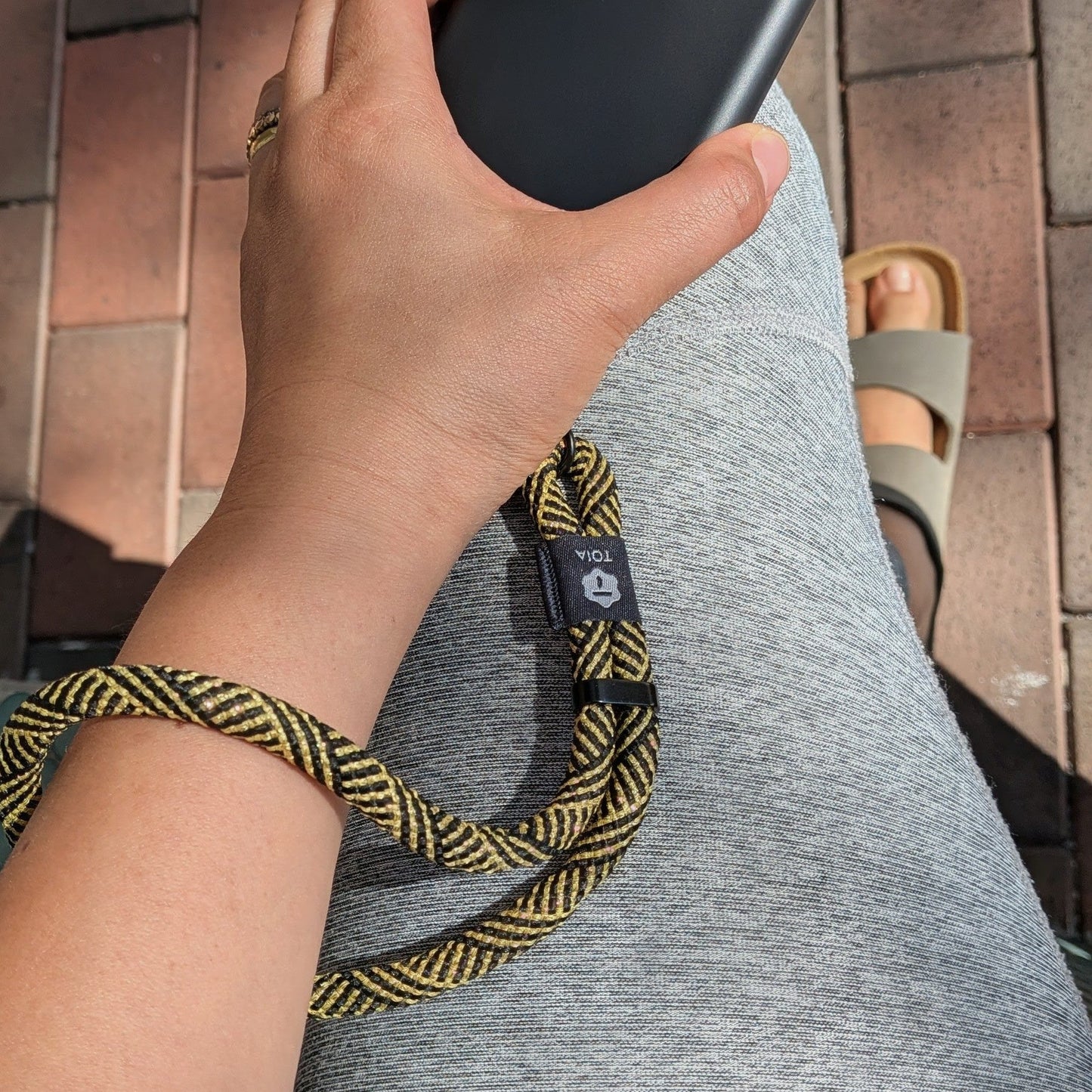 Hand holding a phone attached to a gold and black herringbone patterned wrist phone strap by TOIA.