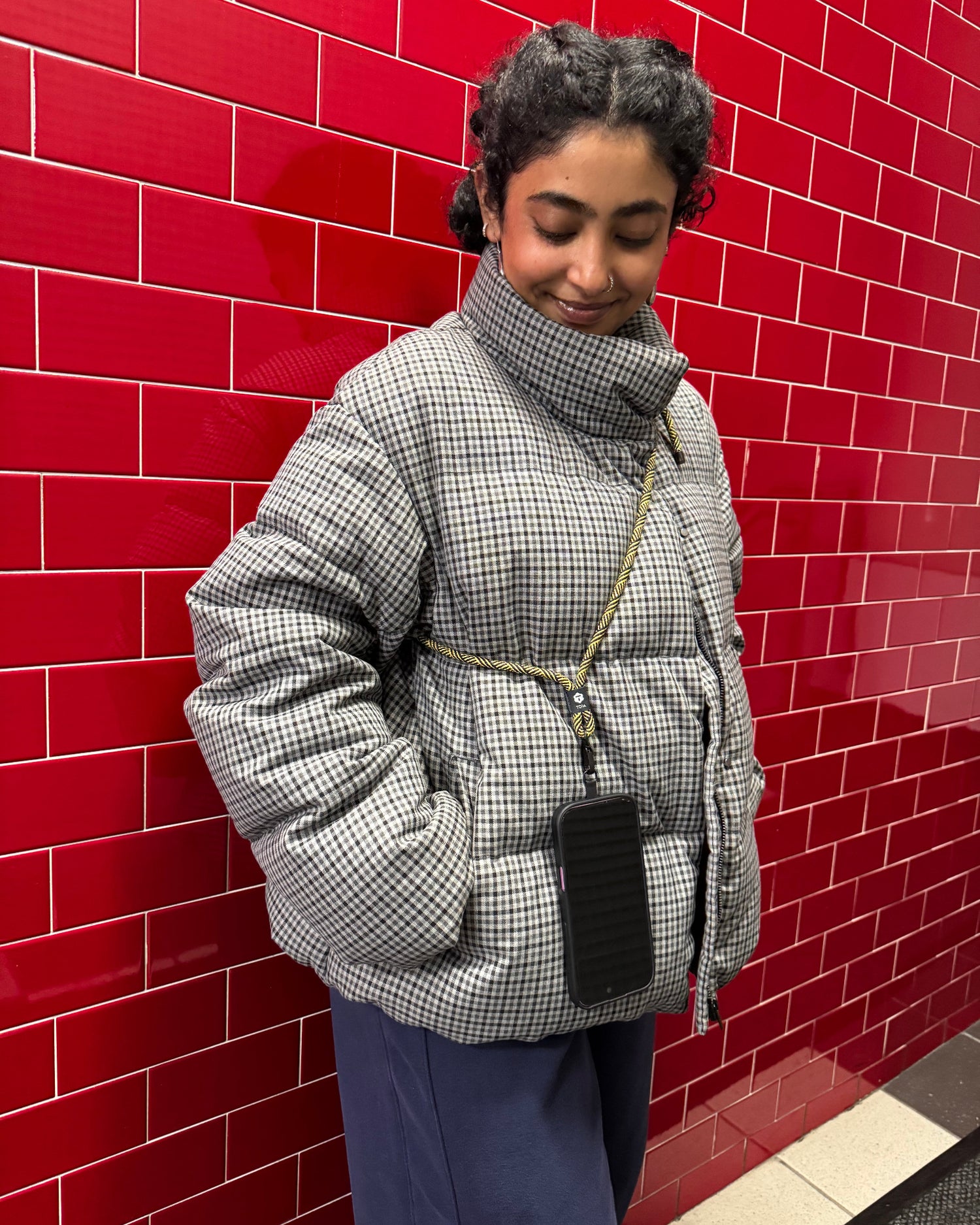Person standing behind on a red tiled wall wearing a checkered coat and toia strap.