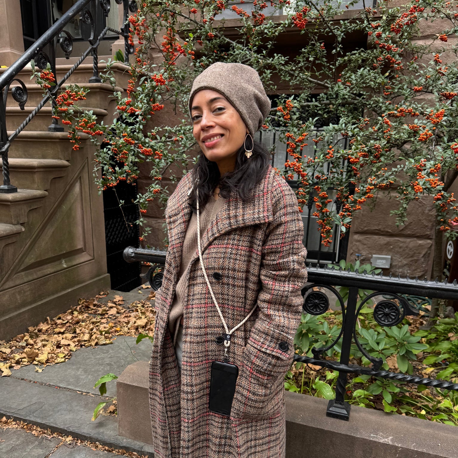 Woman standing in front of Brooklyn brownstone wearing a winter plaid coat with a beige crossbody phone strap by TOIA.
