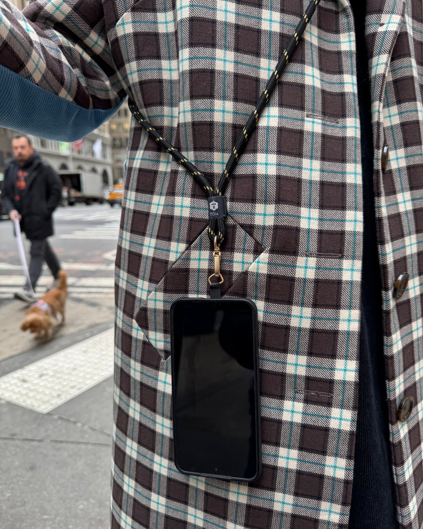 Woman standing on the street wearing a plaid jacket with a smartphone on a black and gold stripe crossbody phone strap by TOIA.