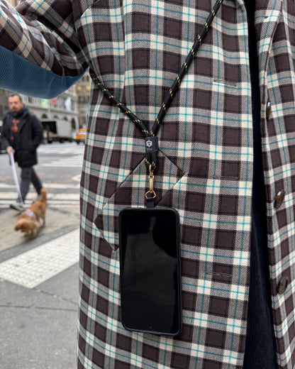 Woman standing on the street wearing a plaid jacket with a smartphone on a black and gold stripe crossbody phone strap by TOIA.