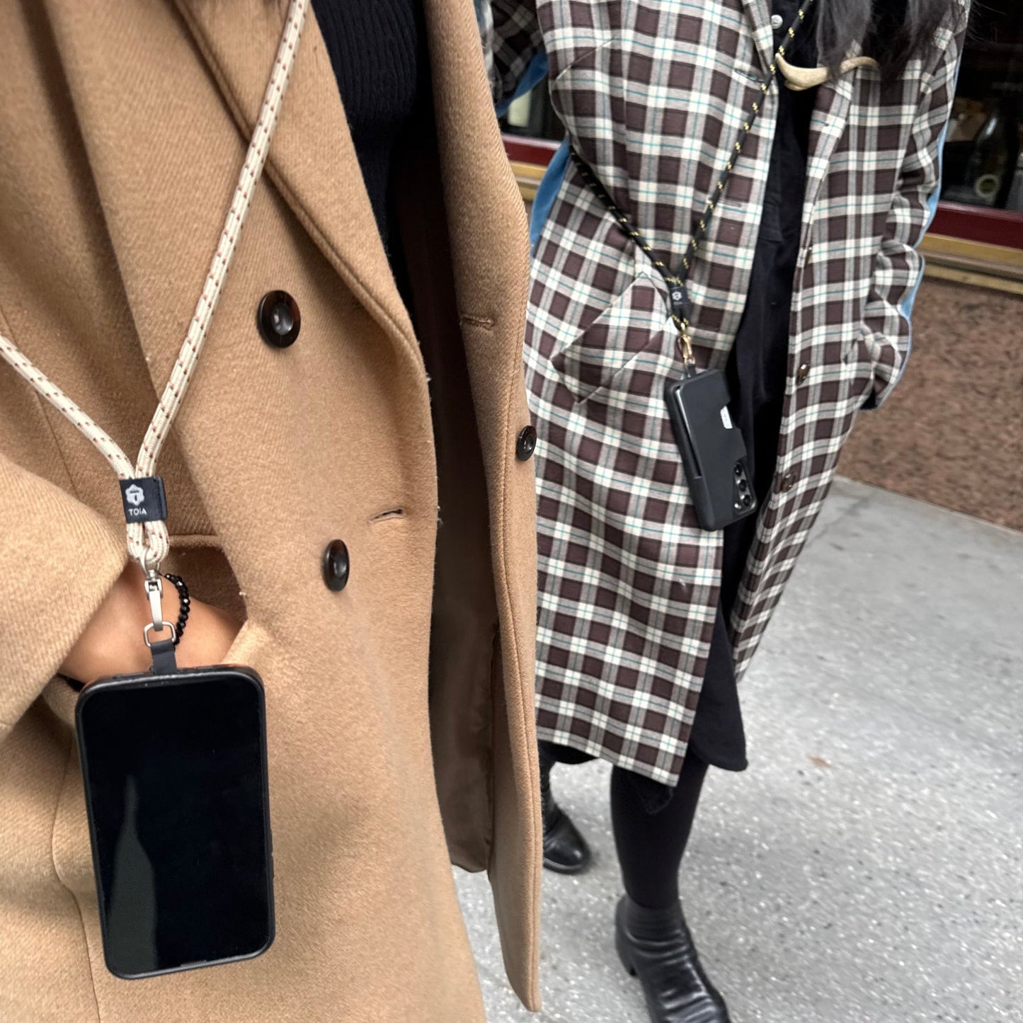 Two women wearing winter coats walking next to each other on a sidewalk carrying smartphones attached to crossbody phone straps by TOIA.