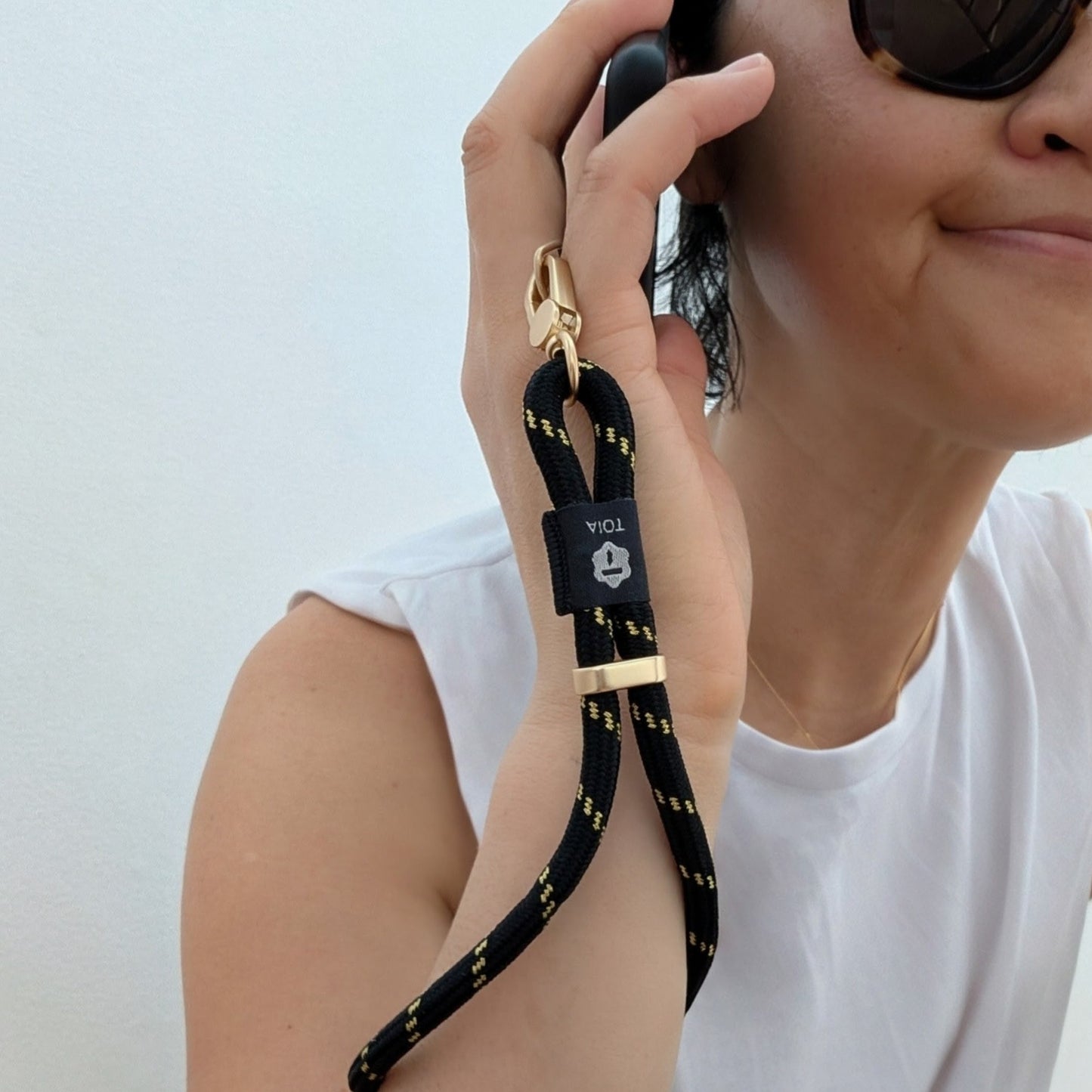 Woman holding her phone to her ear attached to a black and gold stripe phone wrist rope strap with gold hardware and clasp by TOIA.