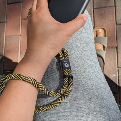 Hand holding a phone attached to a gold and black herringbone patterned wrist phone strap by TOIA. 