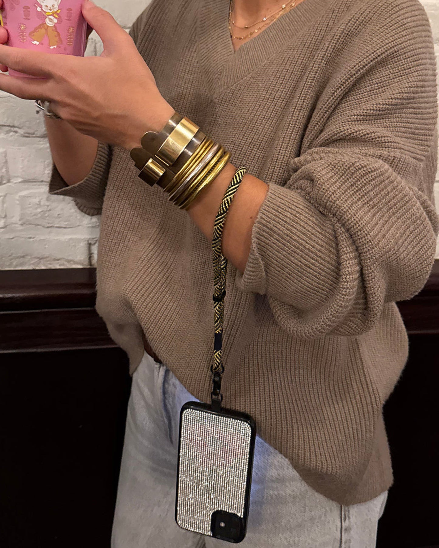 Hand holding a phone attached to a gold and black herringbone patterned wrist phone strap by TOIA. 