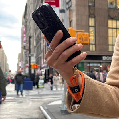 Person holding a phone attached to an orange wrist phone strap by TOIA.