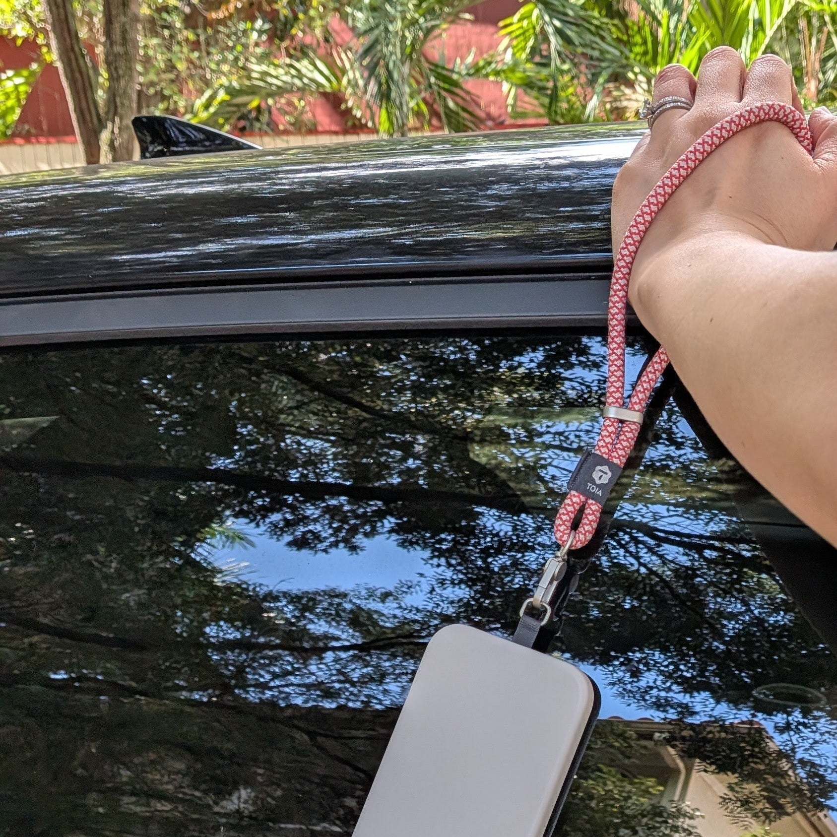 White phone case with a red and white wrist strap attached, held over a car window with trees and a building in the background.